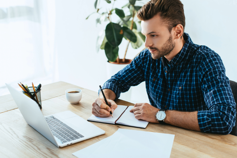 Broker reviewing compliance guidelines on his laptop and taking notes.