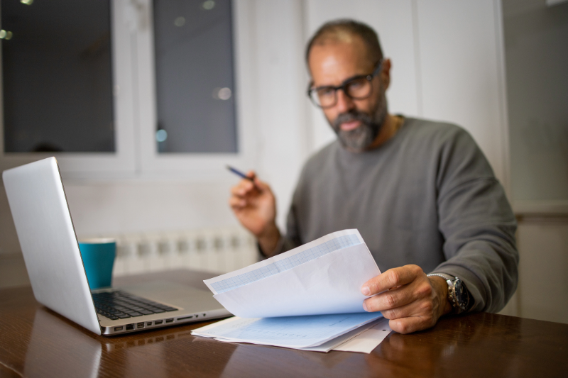 Man with glasses looking at papers with a computer in front of him