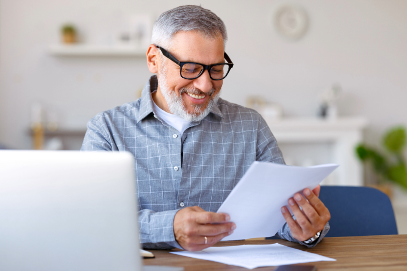 Senior male sitting at a table with a laptop and reviewing paperwork smiling